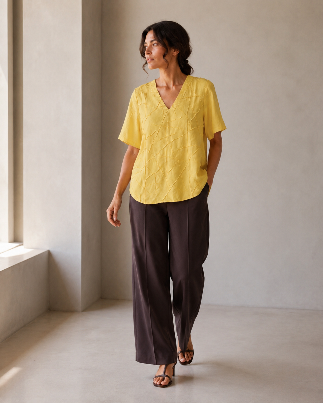 A woman modeling a yellow short-sleeve V-neck blouse with textured embroidery, paired with dark brown wide-leg trousers, standing in a minimalist beige room.