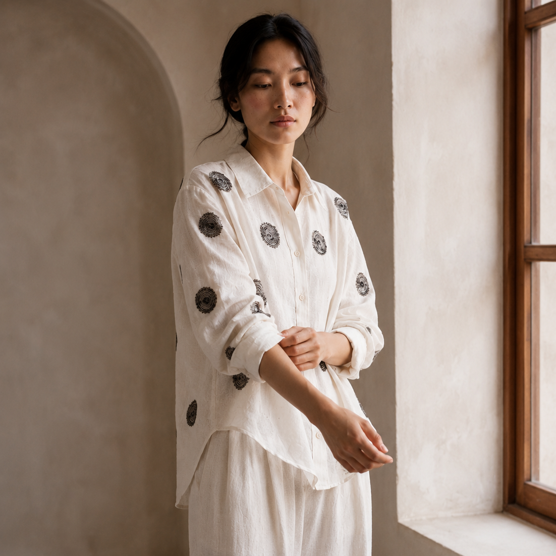 A woman wearing a white long-sleeve button-up shirt with black circular embroidered motifs and matching white trousers, standing in a softly lit room with textured beige walls and a window.