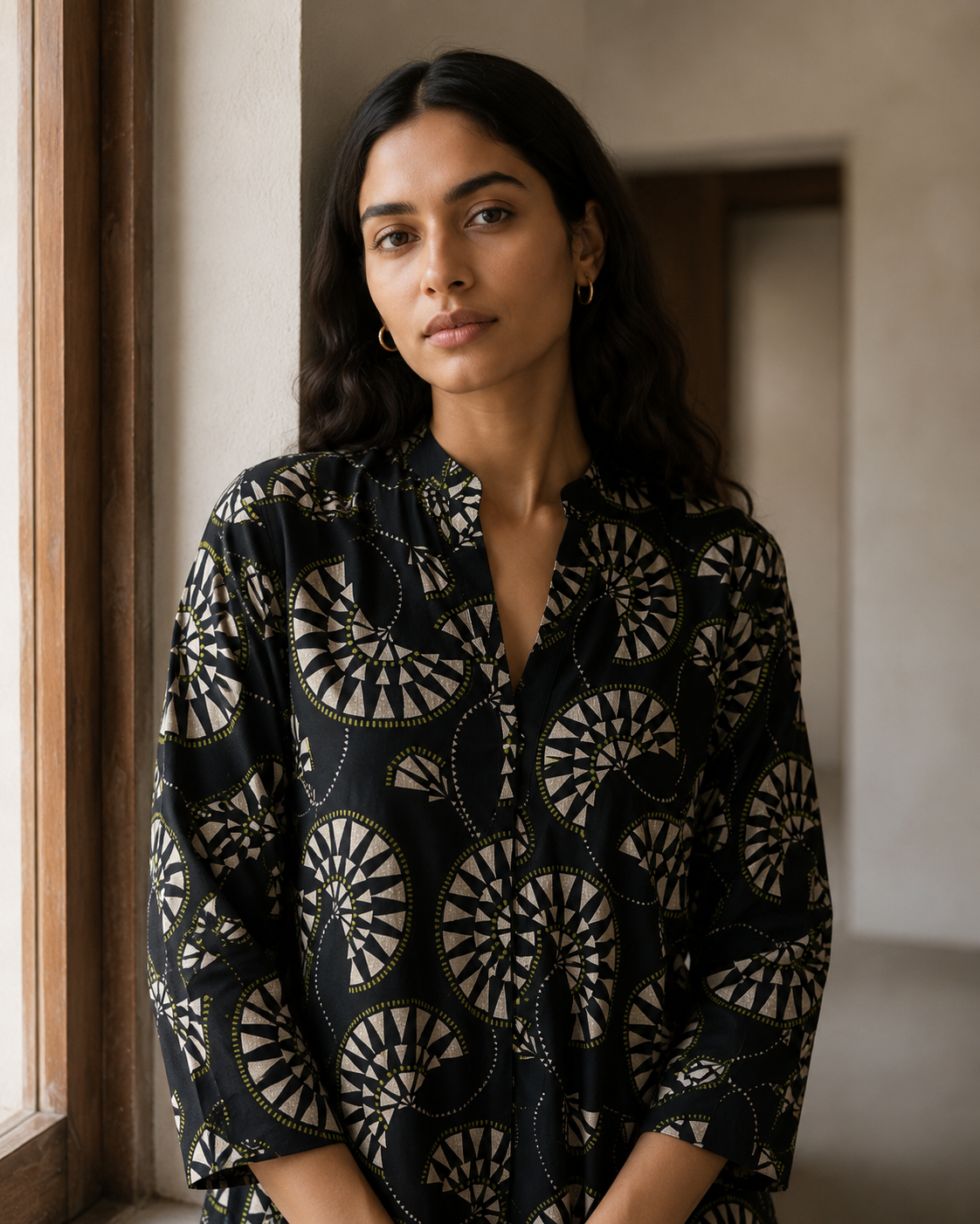 A woman wearing a black box-pleated kurta with a beige-and-white circular geometric pattern, standing in a softly lit indoor setting.